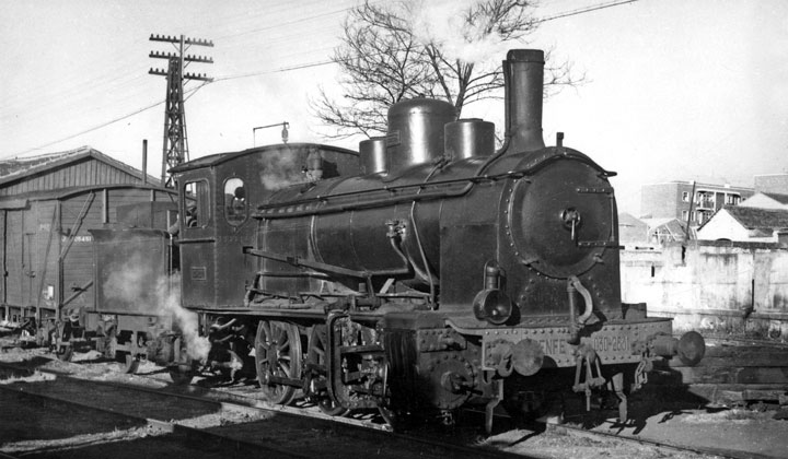 Perspectiva de la locomotora de vapor 030-2531 en Salamanca con un vagón cerrado de mercancías, de madera. Tras ella se ven edificios de viviendas. Archivo Histórico Ferroviario FE-0009-015